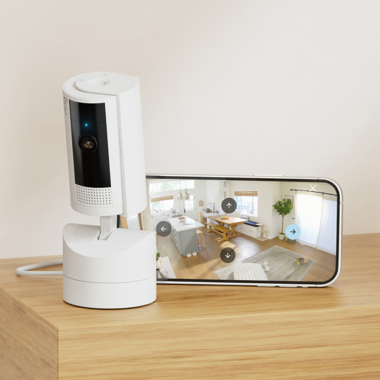 A white security camera sits on a wooden surface next to a smartphone displaying a live video feed of a modern living room.