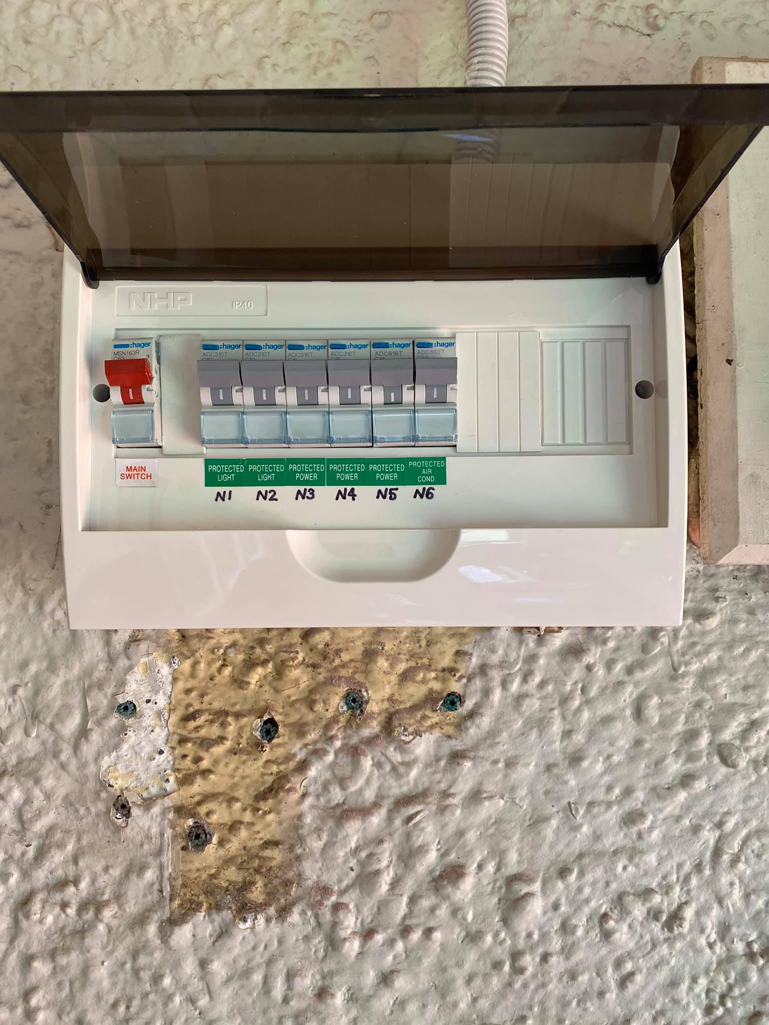 A white electrical breaker panel with labeled switches (N1-N6) mounted on a textured wall with some exposed patches below it.