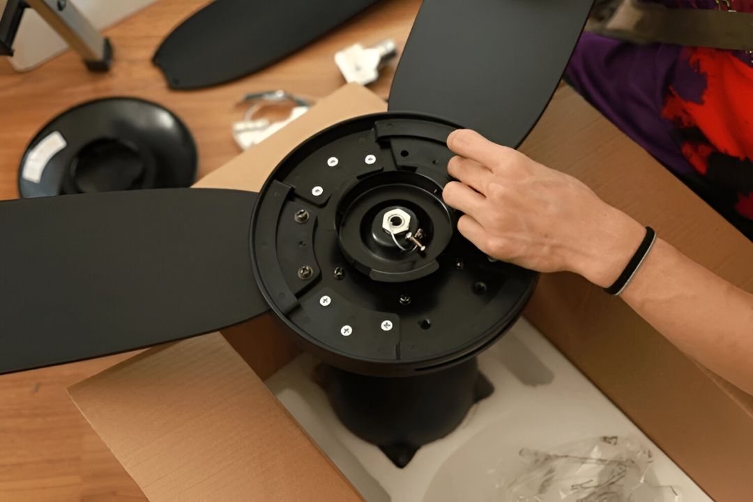 A person assembles a black ceiling fan, securing the blades to the motor housing while working over an open cardboard box, perfect for those tackling ceiling fan repairs in Inner West.