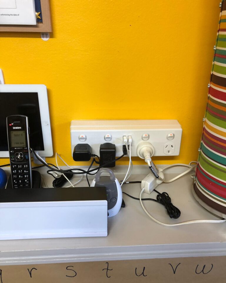A power strip mounted on a yellow wall with multiple devices plugged in, including chargers, a phone, and a baby monitor perfect for showcasing efficient USB & Smart Power Points Installation.