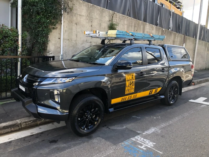 A dark grey utility pickup with ladders and Inner West Electrical Solutions sign is parked on a city street.