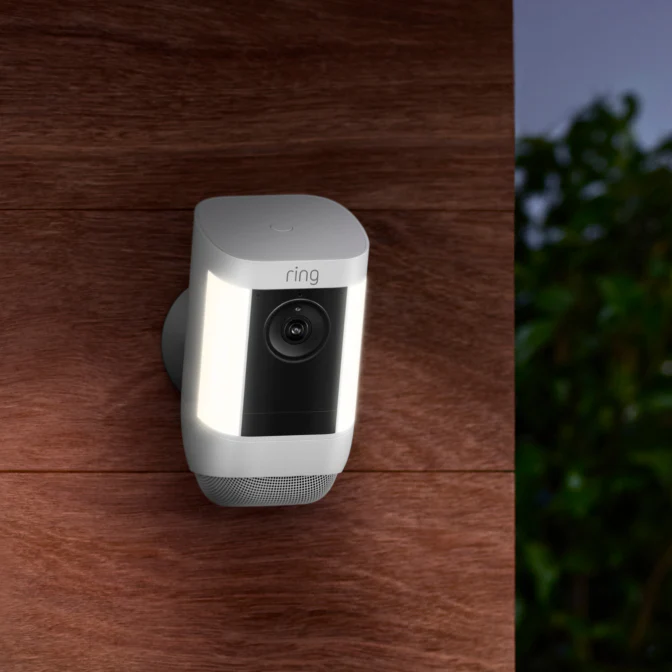 A Ring Security Camera with built-in light is mounted on a Ashfield wall, with lush greenery visible in the background.