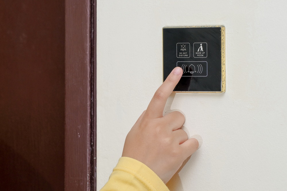 Hand pressing the doorbell button on a wall-mounted electronic room control panel featuring various service icons.
